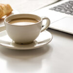 Eine Tasse Kaffee und leckere Croissants, stehen auf dem Schreibtisch neben einem aufgeklappten Laptop. Es geht um eine bezahlte Frühstückspause.
