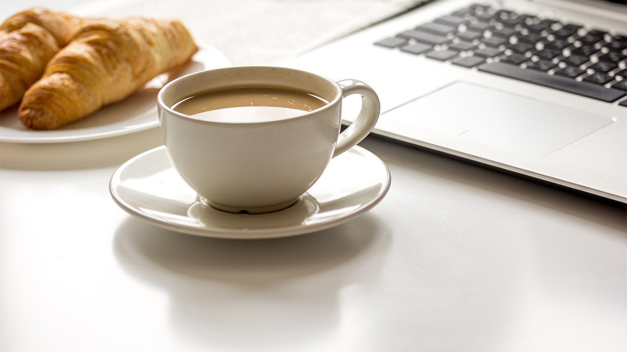 Eine Tasse Kaffee und leckere Croissants, stehen auf dem Schreibtisch neben einem aufgeklappten Laptop. Es geht um eine bezahlte Frühstückspause.