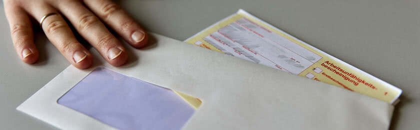 Eine Hand schiebt eine Briefumschlag über den Tisch. Dieser enthält die Arbeitsunfähigkeitsbescheinigung. Der Beschäftigte erhält daraufhin eine Kündigung wegen Krankheit.