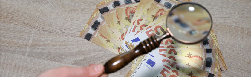 Eine Hand hält eine Lupe über mehrere 50€ Geldscheine und prüft wann ein Anspruch auf eine Abfindung besteht.